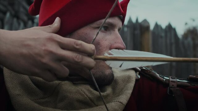 Medium side close-up of young Caucasian medieval archer in red hat drawing bow and aiming feathered arrow at faraway target, while standing guard by wall of wooden military garrison