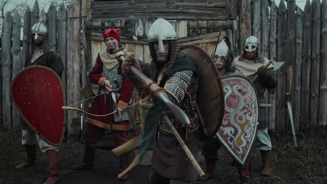 Medium portrait of brave young Caucasian male soldier in helmet, armour aiming lance and looking at camera, while posing with squadron of warriors with swords, shields by gate of wooden garrison