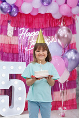 Girl in a hat and with a cake in her hands on the background of a bright photozone and balloons.