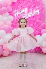 Fototapeta premium Small girl on the background of a photo zone made of pink organza with a neon sign and balloons.