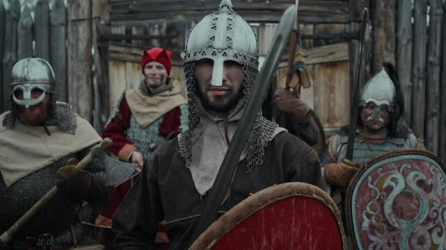 Medium portrait of young bearded Caucasian guard commander in chainmail helmet holding up shield and sword, looking at camera, while standing in front of troop of warriors, ready to defend castle