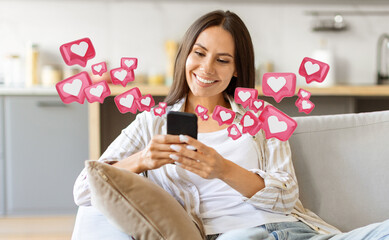 A woman with long brown hair is sitting on a couch in her home. She is smiling as she looks at her smartphone, which is displaying a blank screen. Around her, a number of pink heart icons