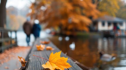 This image features a close-up of an autumn leaf resting on a wooden railing beside a lake, with two people walking in the blurred background, capturing a serene fall moment.