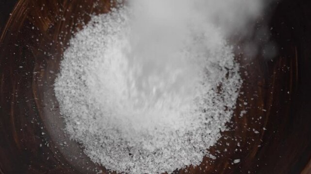 White magnesium salt for relaxing spa falls into a dark wooden bowl creating a stream of sparkling particles in slow motion close up. Rotation