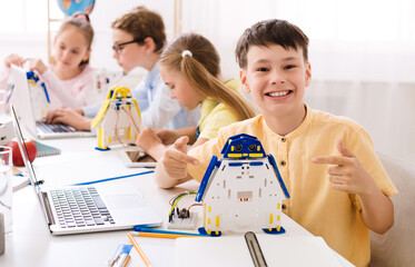 A young boy sits in a classroom, beaming with pride as he points to a robot he built. He is surrounded by other students who are also engaged in STEM activities.