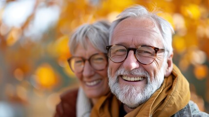 A senior man and woman, both with white hair and glasses, warmly smile at the camera, standing close together outdoors with vibrant autumn leaves in the background.