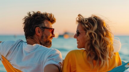 A young couple sits close together in a hammock by the beach, relaxed and enjoying the colorful sunset while the serene ocean landscape stretches out before them.