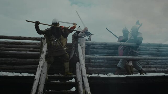 Full low-angle shot of two groups of male soldiers clad in armor, helmets fighting with swords, shields, axe, spear on battlements of wooden military fortress, in billowing smoke, during enemy raid