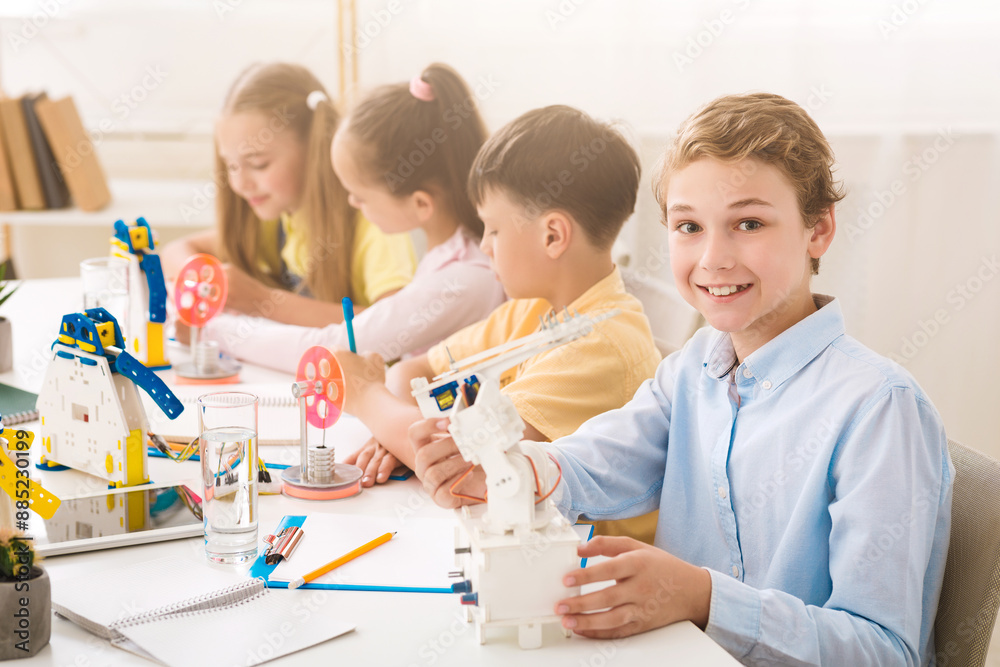 Poster A young boy smiles at the camera while holding a robot during a STEM class. He is sitting at a table with other children who are also working on STEM projects. The classroom is bright and airy