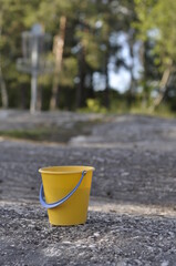 Closeup of bright yellow plastic bucket. Round bucket left behind on a mossy granit rock. Colorful object in focus and green forest in background.