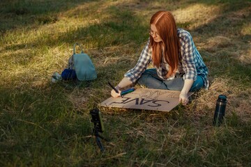 Redhead teenage girl making protest banner and vlogging through smart phone sitting on grass at park