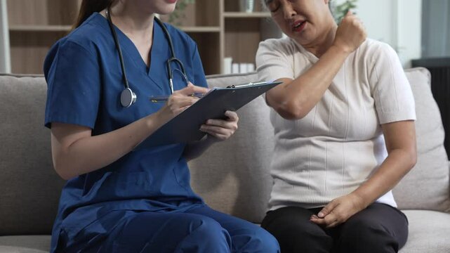 Young Asian nurse attends to older grey-haired mature woman sitting on sofa, conducting health assessment and discussing conditions osteoporosis, arthritis, Alzheimer's disease, dementia, and more.