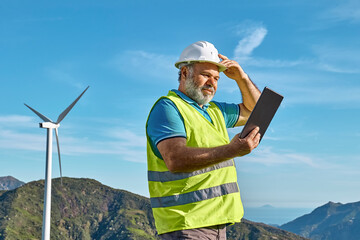 Smiling senior engineer wearing hardhat and using tablet PC in front of wind turbine on sunny day