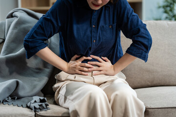 A young Asian woman sits on a sofa in her living room, feeling sick with a stomach ache. She experiences nausea, bloating, and cramping, seeking comfort and relief at home.