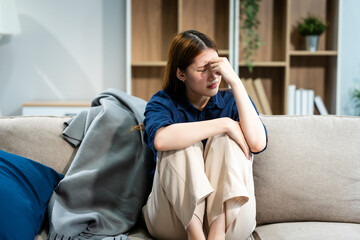 A young Asian woman sits on a sofa in her living room, feeling sick with mental health diseases. She struggles with depression, anxiety, and other conditions, seeking support and solace at home.