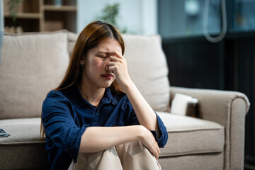 A young Asian woman sits on a sofa in her living room, feeling sick with mental health diseases....