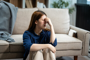 A young Asian woman sits on a sofa in her living room, feeling sick with mental health diseases. She struggles with depression, anxiety, and other conditions, seeking support and solace at home.