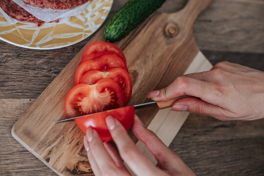 Hands of woman slicing tomato with knife on cutting board in kitchen