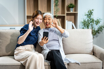 Obraz premium A mature mom and a young Asian woman, mother and daughter, sit together on a sofa in their living room, celebrating Mother's Day with love and bonding at home.