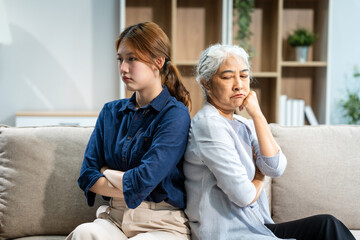 A mature mom and a young Asian woman, mother and daughter, sit together on a sofa in their living...