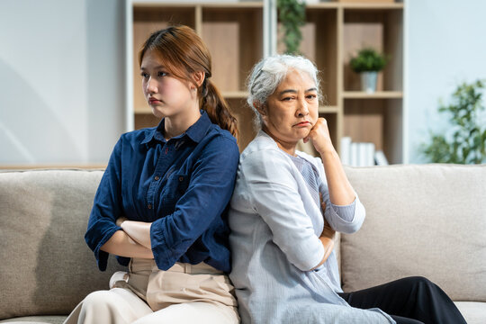 A mature mom and a young Asian woman, mother and daughter, sit together on a sofa in their living room, dealing with an argument and conflict, trying to resolve their differences at home.
