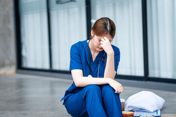 A young Asian woman, possibly a nurse, sits outdoors in front of a hospital, appearing tired and...