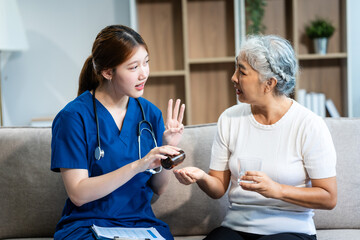 Young Asian nurse attends to older grey-haired mature woman sitting on sofa, conducting health...