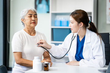 Fototapeta premium A young female nurse works at a desk with a grey-haired Asian mature woman, discussing heart-related conditions like heart disease, heart attack, and hypertension.