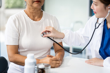 A young female nurse works at a desk with a grey-haired Asian mature woman, discussing heart-related conditions like heart disease, heart attack, and hypertension.