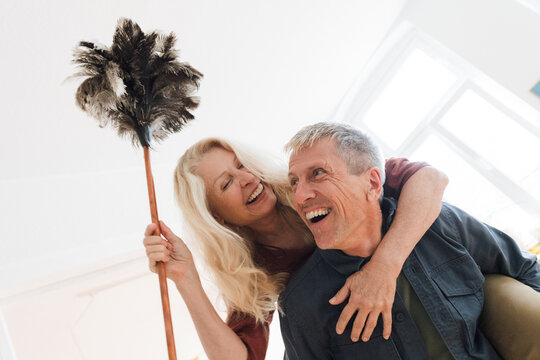 Happy senior man piggybacking blond woman with duster at home
