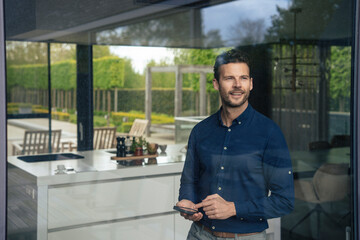 Smiling businessman standing with smart phone seen through window