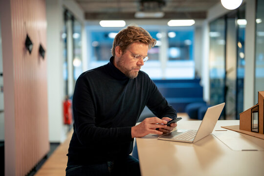 Mature businessman using smart phone and sitting with laptop at desk in office