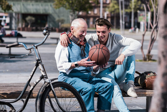 Smiling young man with grandfather holding basketball and sitting on seat at street