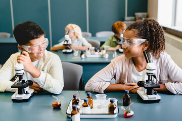 Two mixed-race multi-ethnic classmates kids schoolchildren listening to the chemistry science class lesson at school lab wearing protective eyeglasses using reagents.