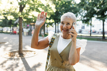 Obraz premium Outdoor portrait of happy pretty elegant caucasian female of 60s in elegant dress calling to her friend to find her in public park and greeting her bestie waving hand, walking on hot summer day