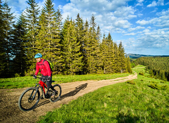 Obraz premium Cyclist man riding electric mountain bike outdoors on sunny day. Male tourist biking along grassy trail in the mountains, wearing helmet and backpack. Concept of sport, active leisure and nature.