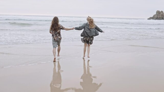 Joyful Gen Z couple enjoys fun summer evening at the beach, running, walking on sand in the ocean. Embracing love, happiness, smile, savor vacation together on the coast, creating lasting memories.