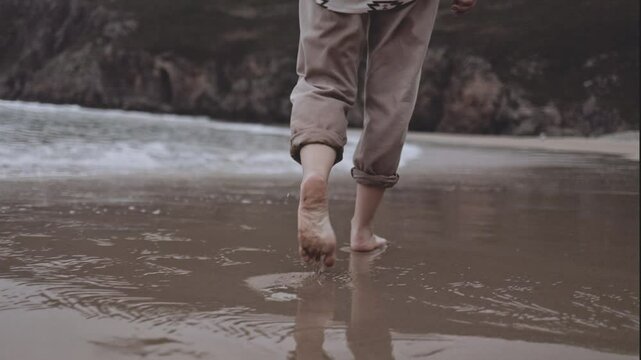 An unrecognizable woman walks alone on tranquil beach at sunset. The calm ocean waves wet the sand and her feet. The cinematic scene captures the melancholy and reflective mood of a summer