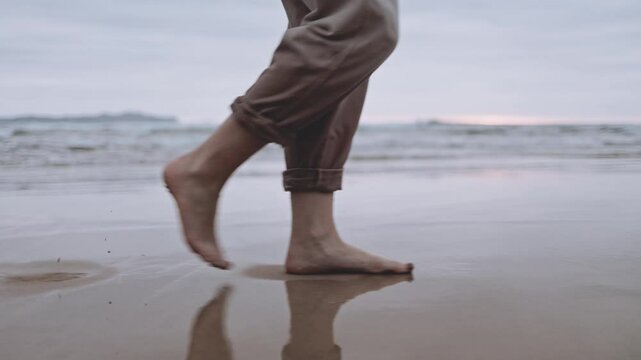 An unrecognizable woman walks alone on tranquil beach at sunset. The calm ocean waves wet the sand and her feet. The cinematic scene captures the melancholy and reflective mood of a summer