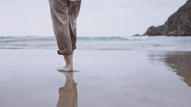 An unrecognizable woman walks alone on tranquil beach at sunset. The calm ocean waves wet the sand and her feet. The cinematic scene captures the melancholy and reflective mood of a summer