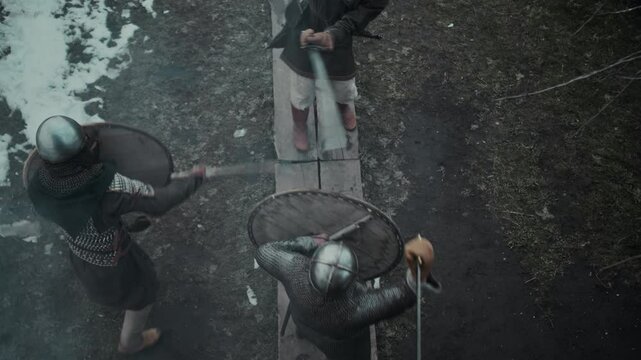 Top shot of skilled ambidextrous male knight in helmet, with two swords fighting alone against attacking enemies with shields and axes, retreating on wooden walkway while defending medieval fortress