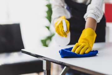 A young woman wearing an apron is cleaning a table in her home office using disinfectant and a wipe.