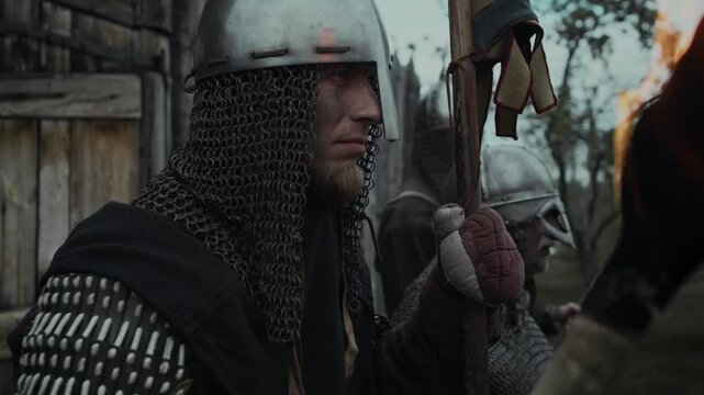 Medium close-up side shot of group of medieval warriors in helmets, holding spear and burning torch, standing by wooden fence on watch duty, peering away at distance and talking