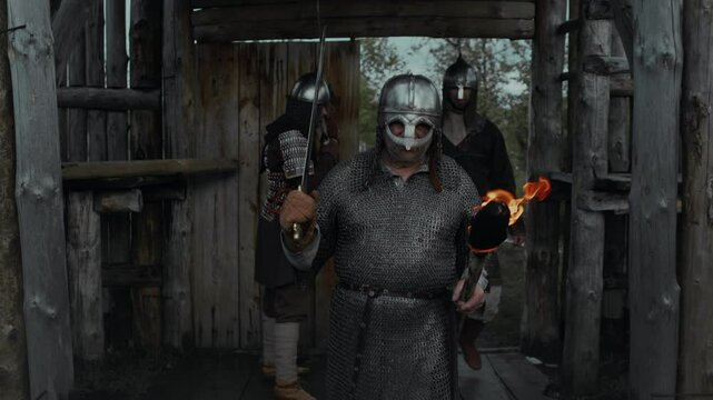 Medium full tracking shot of feudal castle guard in armoured tunic and helmet opening wooden gate to let in comrades returning from patrol, walking and carrying swords and burning torches