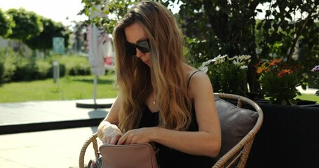 Stylish woman with loose hair seeks cosmetics in designer handbag in summer park. Female with sunglasses sits in wicker armchair on cafe terrace
