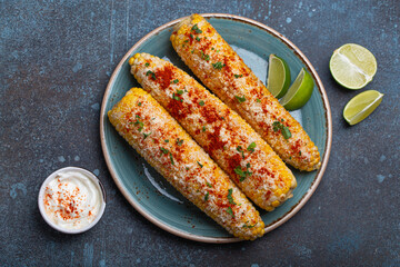 Three grilled corn cobs coated in spices, mayonnaise, and cotija cheese are served on a plate with lime wedges and chili powder sour cream