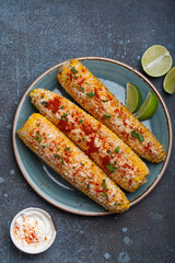 Three grilled corn cobs coated in spices, mayonnaise, and cotija cheese are served on a plate with lime wedges and chili powder sour cream