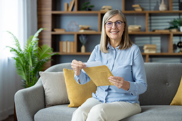 Happy senior woman sitting on grey sofa in cozy living room opening a letter with joy. She is...