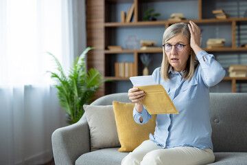 Shocked elderly woman receiving unexpected news while reading letter on sofa at home. Senior lady...