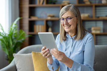 Smiling senior woman using digital tablet at home, sitting on sofa. Enjoying technology, staying connected with family and friends. Modern living room with natural light, bookshelf, and green plant.